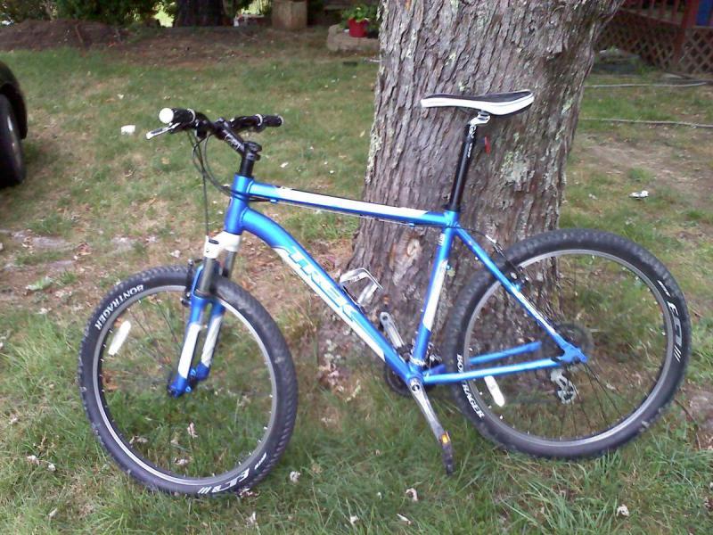 Trek 3700: A blue mountain bike leaning against a tree, set on a grassy area. The bike features thick tires, a dual suspension system, and a sleek frame, with a visible seat and handlebars. In the background, there is a partially visible black car and greenery.