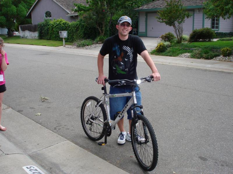 Trek 3700: A smiling young man in a black t-shirt and denim shorts stands next to a silver and black mountain bike on a residential street. A young girl in a pink shirt is standing nearby, appearing to be observing him. Lush greenery and houses can be seen in the background.