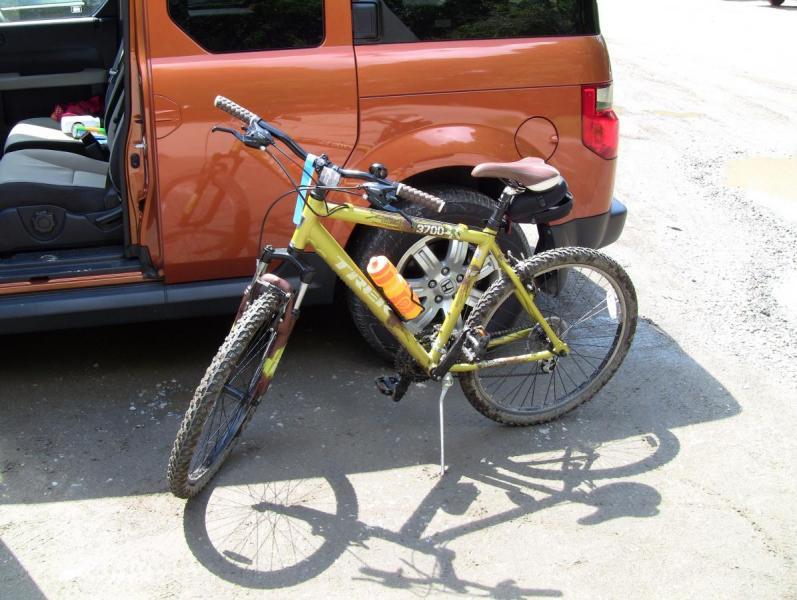 Trek 3700: A parked mountain bike with yellow accents stands beside an orange vehicle. The bike features a water bottle holder with an orange bottle and is positioned on a gravel surface, with the door of the vehicle open, showing items inside.