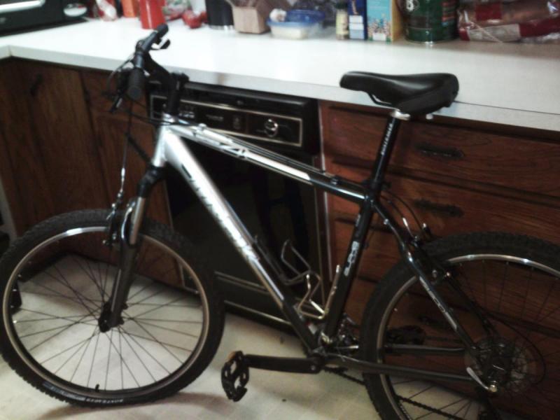 Trek 4300: A silver and black mountain bike is leaning against a kitchen counter, showcasing its sturdy frame and thick tires. The background features wooden cabinets and various kitchen items, indicating a homely setting. The bike is positioned in a way that highlights its handlebars and seat.