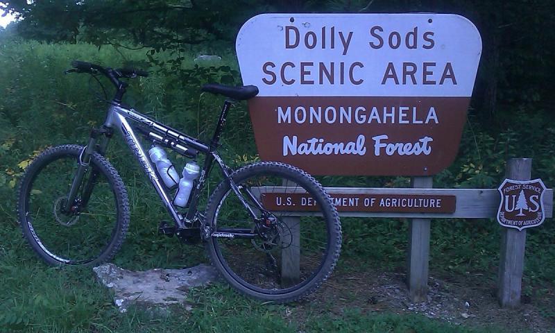 Trek 4300: A mountain bike leaning against a sign that reads "Dolly Sods Scenic Area, Monongahela National Forest." The sign is brown and white, indicating the entrance to the scenic area. Green foliage and trees are visible in the background.