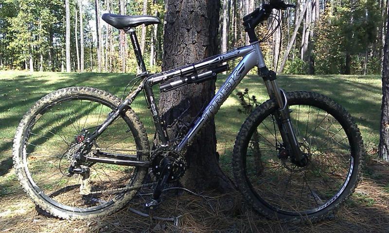 Trek 4300: A silver Trek mountain bike leaning against a tree, surrounded by a grassy area and trees in the background. The bike has visible mud on the tires and frame, indicating recent use on trails.