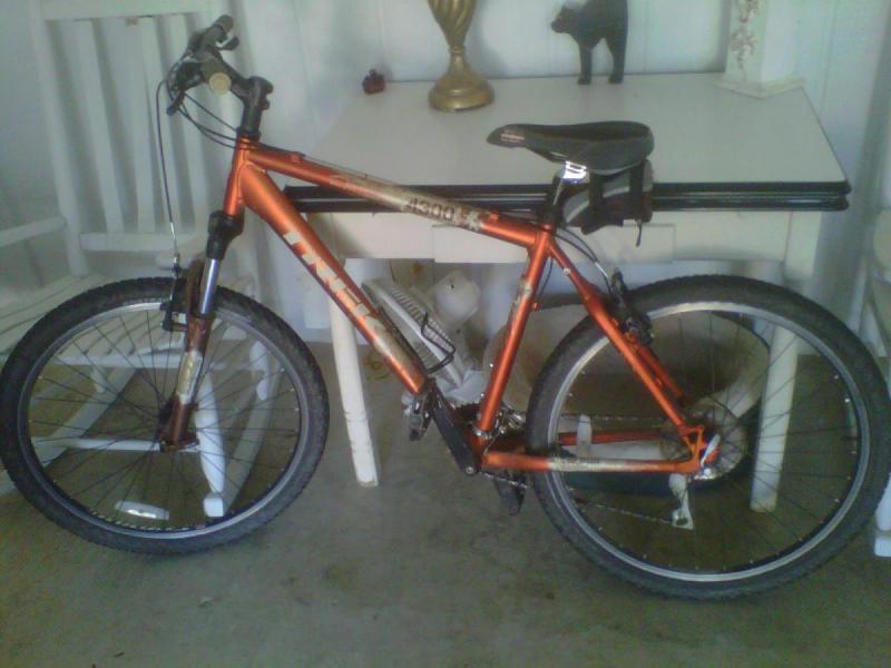 Trek 4300: A mountain bike with an orange frame, black handlebars, and chunky tires is positioned next to a small table. The background includes a white chair and a decorative object on the table. The bike has a water bottle holder and a seat bag attached.