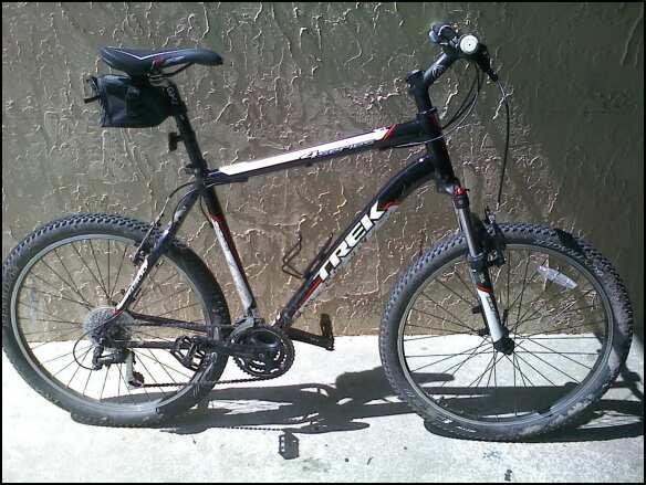 Trek 4300: A black and silver Trek mountain bike is leaning against a textured wall. The bike features wide tires, a front suspension, and a small black bag attached to the back of the seat. Sunlight is casting shadows on the ground, highlighting the bike's details.
