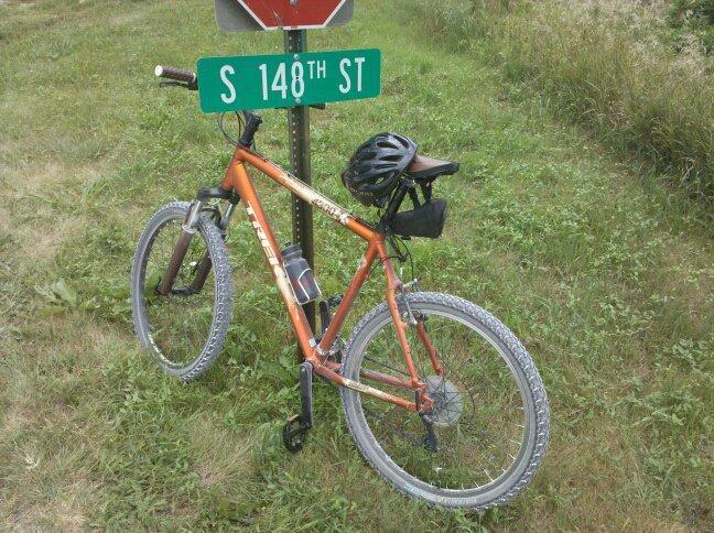 Trek 4300: A mountain bike with an orange frame is leaning against a green street sign that reads "S 148th ST." The bike is equipped with a black helmet on the handlebars and a small bag attached to the frame. The background features a grassy area, suggesting a rural or suburban setting.