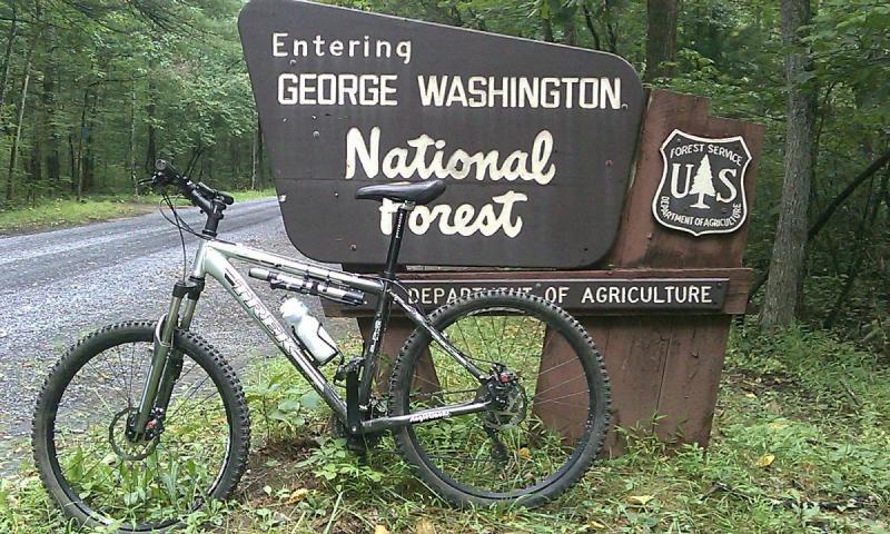 Trek 4300: A mountain bike parked next to the entrance sign for George Washington National Forest, surrounded by trees and a gravel road. The sign displays "Entering George Washington National Forest" and includes the U.S. Forest Service logo.