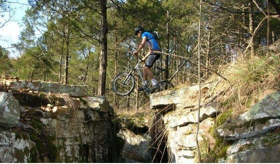 Trek 4300: A mountain biker navigating a narrow stone ledge over a rocky gap in a forested area, with trees and underbrush surrounding the scene.