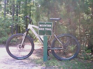 Trek 4300: A mountain bike leaning against a sign that reads "MOUNTAIN BIKE TRAIL," surrounded by a forested area with trees and foliage.