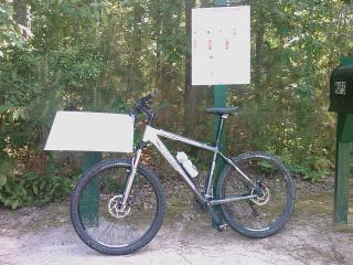 Trek 4300: A mountain bike is parked next to a sign in a wooded area. The sign features a map or information about the surrounding trails, and there is a green mailbox nearby.
