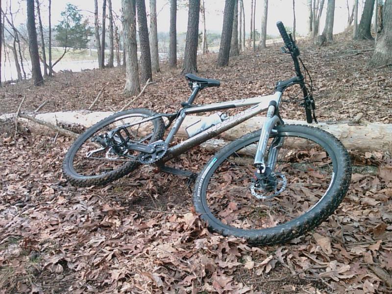 Trek 4300: A mountain bike resting on a fallen log in a wooded area, surrounded by fallen leaves and trees. The bike has a sturdy frame and is partially covered in mud, indicating recent use on rough terrain. In the background, a glimpse of water can be seen through the trees.