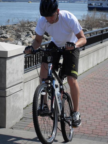 Trek 4300: A young man wearing a white t-shirt and black shorts rides a mountain bike, balancing on the front wheel with one foot off the ground. He is in an outdoor setting with a path beside a rocky shoreline and water in the background. The scene is bright and sunny.