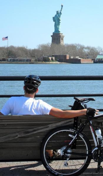 Trek 4300: A person in a white t-shirt and bike helmet sits on a wooden bench, looking out at the Statue of Liberty across the water. A bicycle leans against the bench beside them. In the background, a clear blue sky frames the iconic statue and American flag.