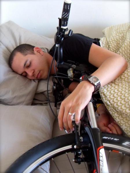 A person sleeping in bed with a bicycle partially propped over them, one arm resting on the bike's wheel and the other arm tucked under a pillow. The scene is cozy and humorous, showcasing a unique interaction between the person and the bicycle.