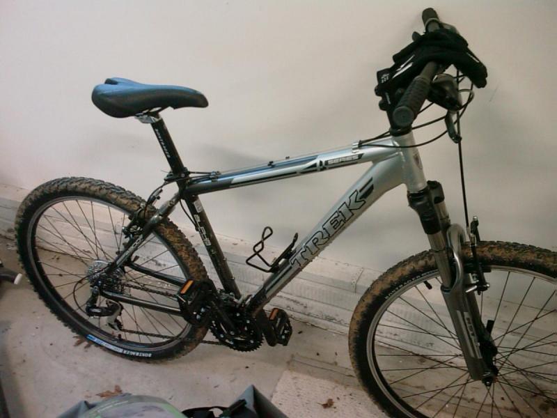 Trek 4300: A silver Trek mountain bike with a black seat, placed against a light-colored wall. The bike features thick, knobby tires, a front suspension fork, and handlebars equipped with grips. It shows signs of use, with some dirt on the tires and frame.