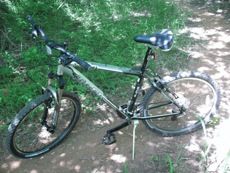 Trek 4300: Mountain bike parked on a dirt trail surrounded by greenery. The bike features a sturdy frame, thick tires, and a saddle, with the Trek brand visible on the frame.