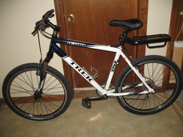 Trek 4300: Alt text: A Trek mountain bike featuring a black and white frame, with a sleek design, wide tires, and a rear rack, positioned in front of a wooden door inside a home.