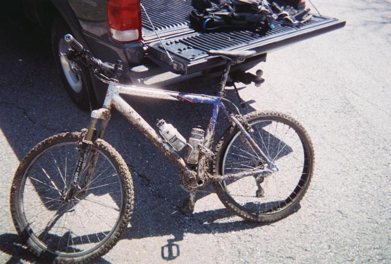 Trek 4300: A muddy mountain bike leaning against a truck tailgate, with two water bottles attached to the frame, parked on a gravel surface. A backpack and other gear are visible on the truck bed in the background.
