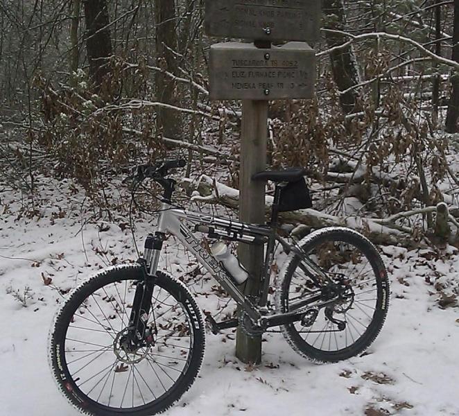Trek 4300: Mountain bike resting on a snowy trail next to a wooden signpost with trail names. The surrounding forest is lightly dusted with snow, and there are bare trees in the background.