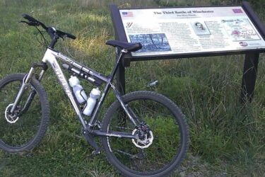 Trek 4300: A mountain bike with water bottles is parked beside an informational sign about the Third Battle of Winchester, set in a grassy area. The sign features historical details and images related to the battle.