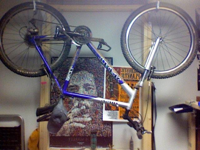 Trek 4500: A mountain bike hanging upside down from hooks in a garage or workshop, with a gray hat resting on the frame. Posters and tools are visible in the background.