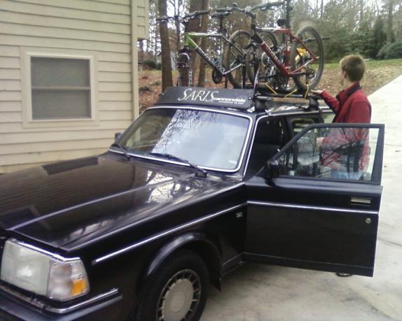Trek 4500: A black car parked in a driveway with a person attaching bicycles to a roof rack. The car's door is open, and there is a residential building in the background among trees.