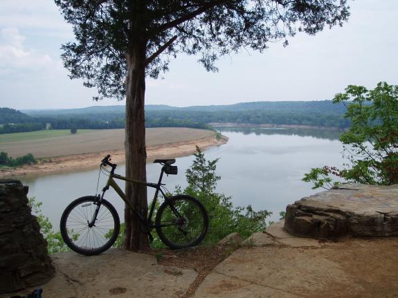 Trek 4500: A scenic view of a river surrounded by green hills, with a bicycle resting against a tree at the edge of a rocky overlook. The sky is partly cloudy, and there are open fields in the background.