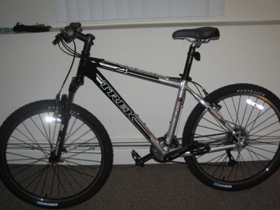 Trek 4500: A black and silver Trek mountain bike is leaned against a wall in a room. The bike features thick, knobby tires and a sturdy frame, with visible gears and handlebars. The setting has a simple interior, with a small window and neutral-colored flooring.