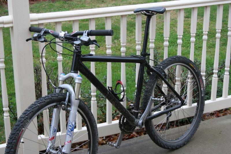 Trek 4900: A black mountain bike with a silver front fork is leaned against a white wooden railing. The bike has thick, knobby tires and a minimalist design, with visible brake and gear cables. The background features green grass and a few decorative shrubs.