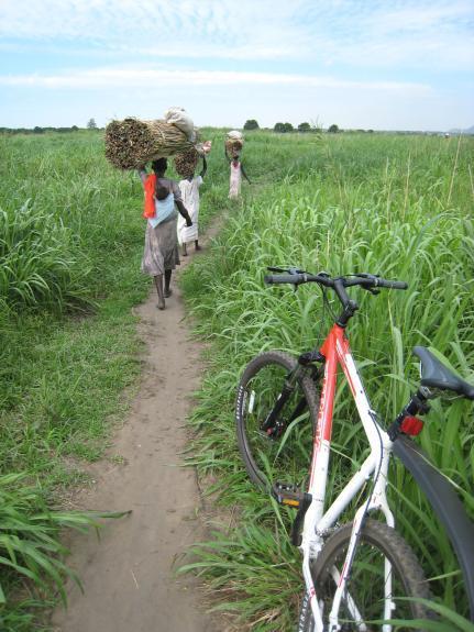 Trek 6000: Three individuals walk along a narrow dirt path through tall green grass, carrying bundles on their heads. A bicycle is parked to the right of the path, partially visible. The scene captures a rural landscape with a clear sky in the background.
