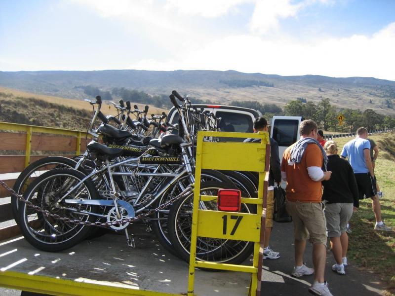 Trek 6000: A group of people gathered near a yellow trailer loaded with several bicycles, set against a backdrop of rolling hills and a partly cloudy sky. Some individuals are interacting and discussing, while others appear to be preparing for an outdoor biking adventure.