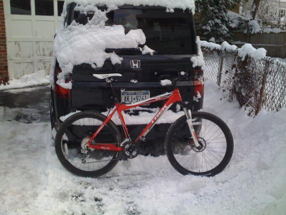 Trek 6000: A red mountain bike is propped against the back of a black SUV, which is covered in snow. The driveway is also filled with a layer of snow, indicating winter weather. A portion of a wooden fence is visible in the background.