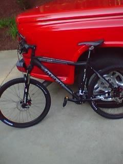 Trek 6000: A black mountain bike with disc brakes parked next to a red pickup truck on a concrete surface. The bike is angled to show its frame and wheels clearly.
