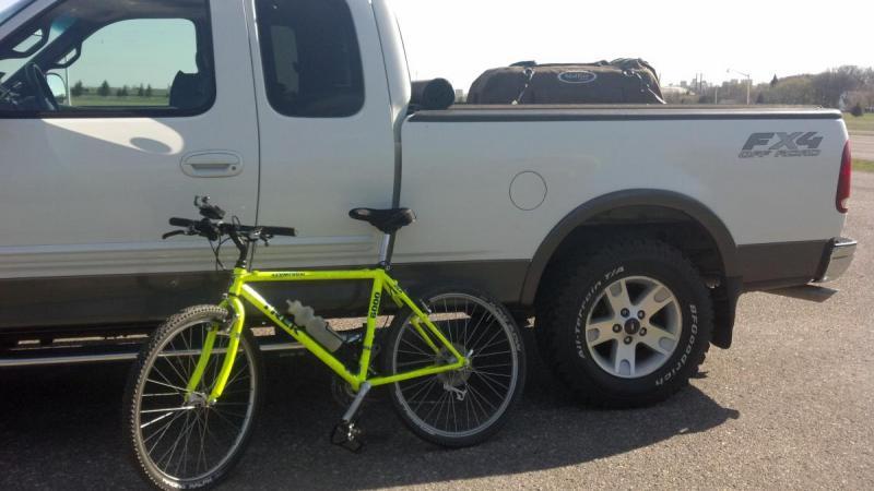 Trek 6000: A bright yellow mountain bike is positioned next to a silver pickup truck parked on a paved surface. The bike stands upright with its front wheel turned, while the truck's bed is loaded with a bag. The setting appears to be outdoors on a clear day.
