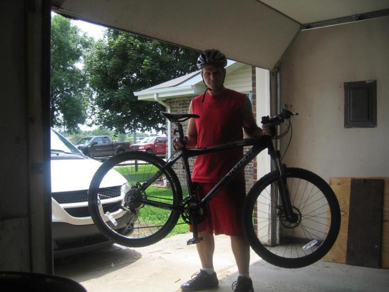 Trek 6000: A person wearing a red sleeveless shirt and a helmet is holding a mountain bike while standing in a garage. The background shows a driveway with a vehicle parked outside and trees in the distance.