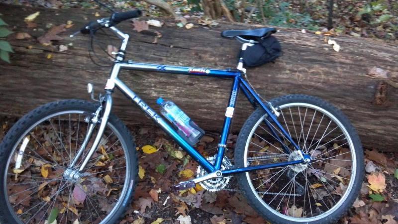 A blue and silver mountain bike leaning against a fallen log, surrounded by autumn leaves. The bike has a water bottle attached to the frame and a small black bag on the seat. The background includes foliage indicating a wooded area.