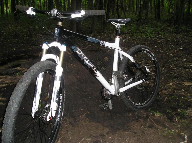Trek 69er: A mountain bike with a black and white frame resting on a dirt path surrounded by greenery, showcasing its front suspension and rugged tires.