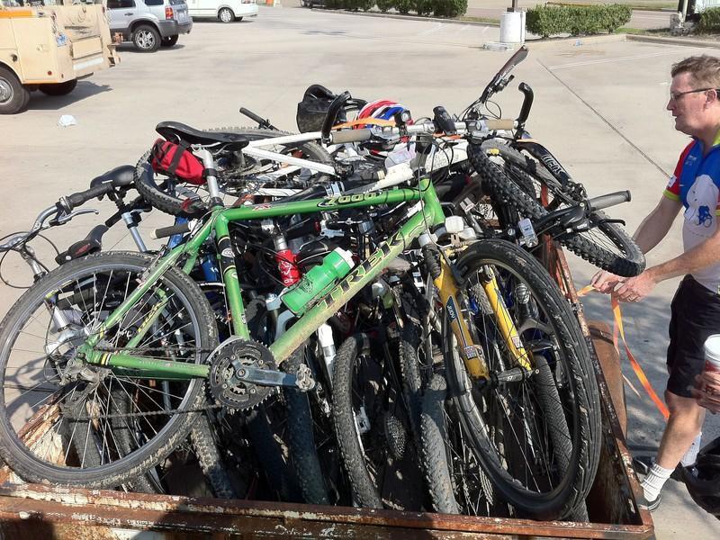 Trek 7000: A pile of bicycles stacked in a storage container, with a man in a colorful cycling jersey inspecting them. Various bikes are visible, including a green Trek bicycle and several others in different colors and styles, set against a parking lot backdrop.