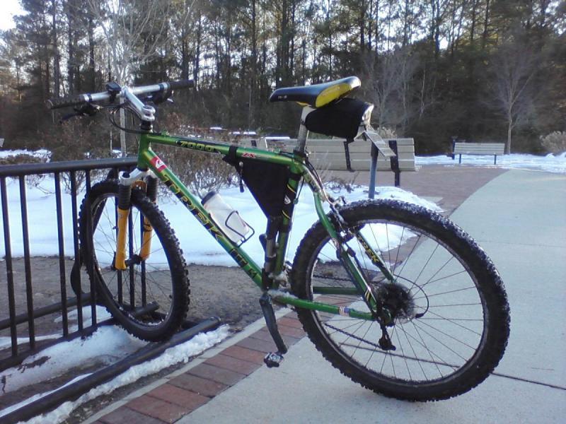 Trek 7000: A green mountain bike with yellow accents is parked against a railing in a park. The ground is partially covered in snow, and benches are visible in the background among trees. The bike features thick tires and a water bottle holder.
