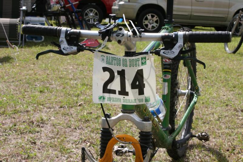 Trek 7000: A close-up view of a mountain bike with a race number 214 attached to the handlebars. The bike is set on a grassy surface, and in the background, there are other bicycles and vehicles. The race number includes the text "SANTOS OR BUST" and a date reference, "SPRING BREAK '06."