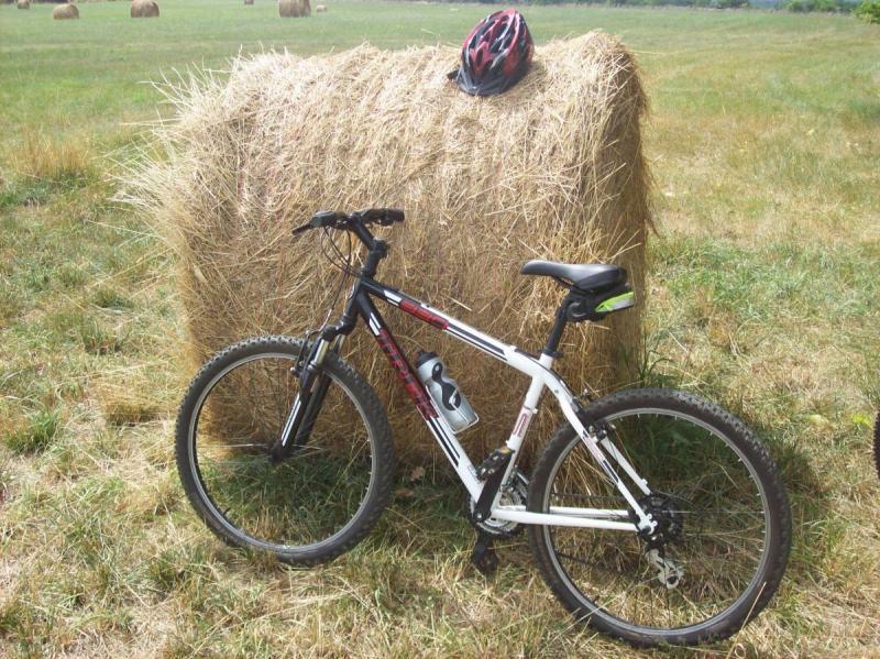 Trek 820: A black and white mountain bike leaning against a round hay bale in a grassy field, with a red bicycle helmet resting on top of the hay bale.