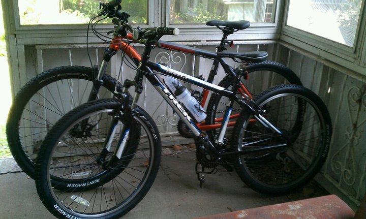 Trek 820: Two mountain bikes parked inside a screened-in porch, one primarily orange and the other black and white. Both bikes have thick tires and a water bottle attached to the frame. The porch features a wooden floor and a view of greenery outside.