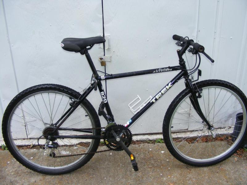 Trek 820: A black Trek 820 mountain bike, featuring a sturdy frame, knobby tires, and a traditional handlebar design, positioned against a white wall. The bike has a comfortable saddle and visible gear components, suggesting it is suitable for off-road riding.
