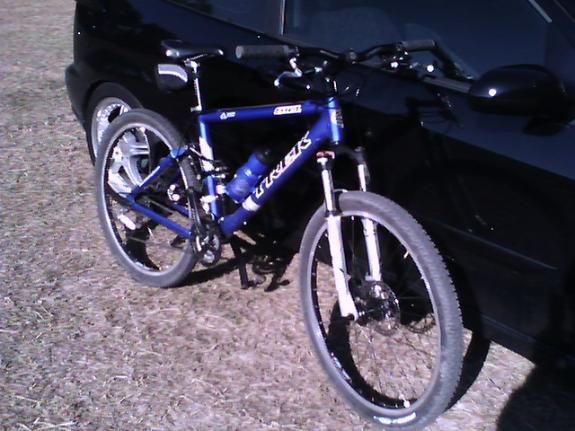 Trek Fuel 70: A blue mountain bike parked beside a black car on grassy ground. The bike features thick tires, a front suspension, and a water bottle holder attached to the frame. Sunlight is shining, creating a clear view of the scene.