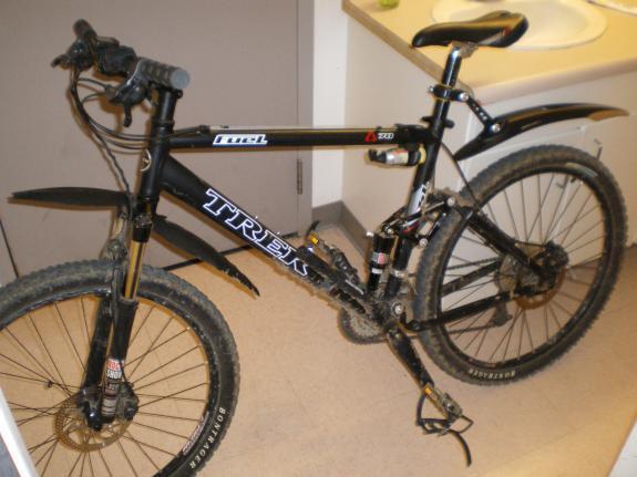 Trek Fuel 70: A black Trek mountain bike with visible dirt on the tires and frame, leaning against a wall in a room with a countertop in the background. The bike features a front suspension fork and has a sleek design, suitable for off-road riding.