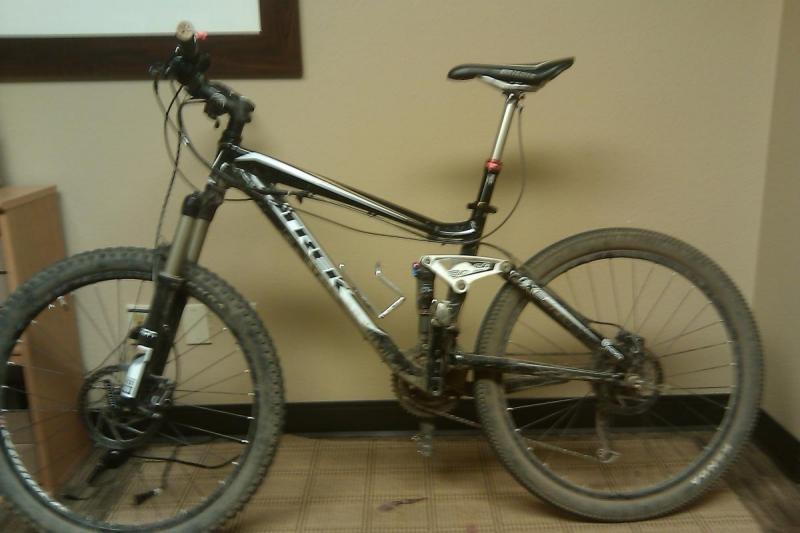 Trek Fuel EX 6: A black and silver mountain bike with thick tires and front suspension, resting against a wall in an indoor setting. The bike appears to have some dirt on it, indicating recent outdoor use. A wooden cabinet is partially visible in the background.