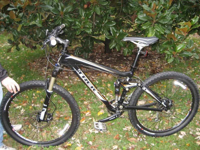 Trek Fuel EX 6: A black Trek mountain bike with front suspension, large tires, and visible gears, leaning on a person's leg, set against a backdrop of green foliage and fallen leaves.