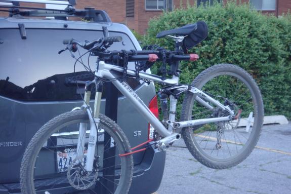 Trek Fuel EX 7: A silver mountain bike is mounted on a bicycle rack attached to the back of a dark SUV. The bike's front wheel is raised, and the rear wheel is secured with straps. The background features greenery and a parking area.