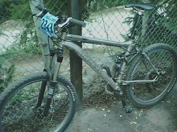 Trek Fuel EX 7: A mountain bike leaning against a wooden fence, featuring a race number tag labeled "2341." The bike has a silver color with visible suspension components and knobby tires, set in a natural outdoor environment with greenery in the background.