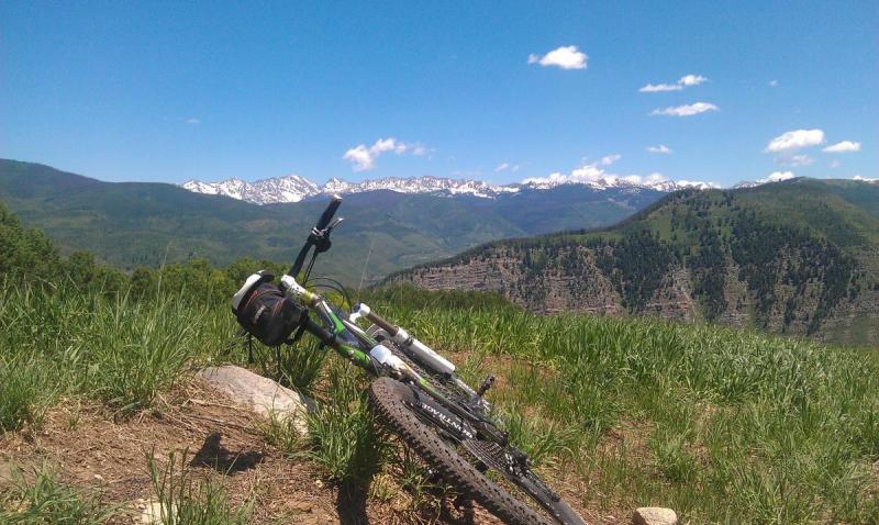 Trek Fuel EX 8: A mountain bike rests on a rocky outcrop surrounded by lush green grass, with a panoramic view of snow-capped mountains in the background under a clear blue sky dotted with a few clouds.