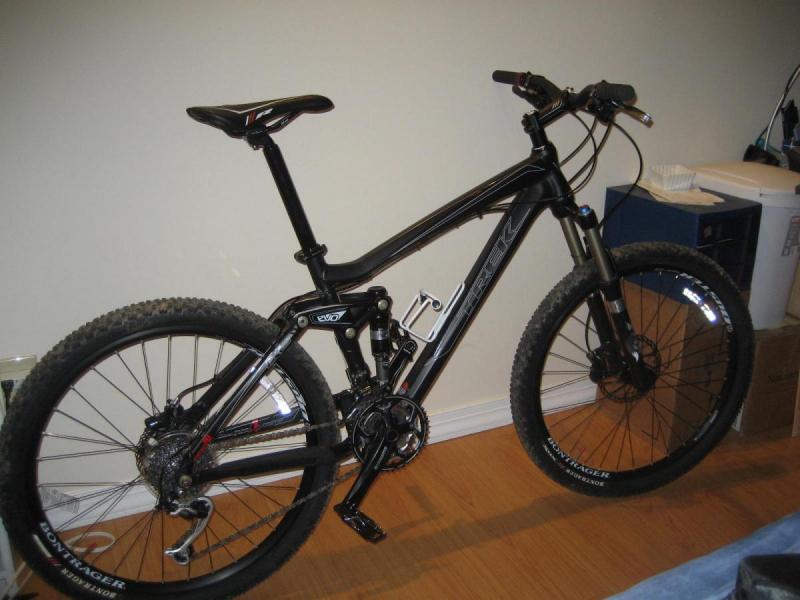 Trek Fuel EX 8: Image of a black mountain bike leaning against a wall inside a room. The bike features thick tires, a dual suspension system, and a sleek frame. The background includes a wooden floor and scattered items like a box and a container.